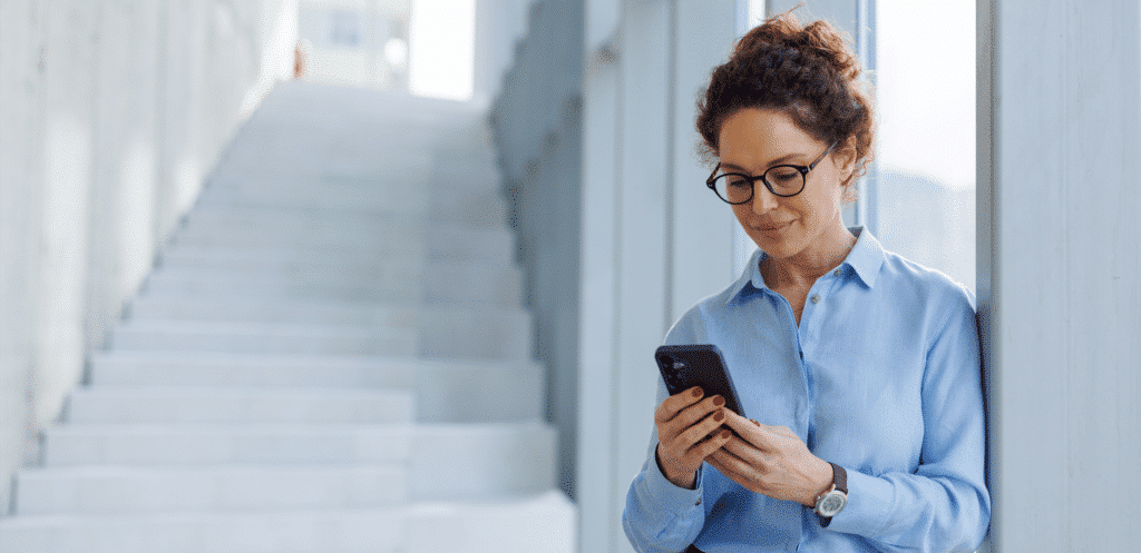 A woman wearing glasses is leaning against a wall in a modern building and looking at her smartphone; in the background, a staircase leads upward.