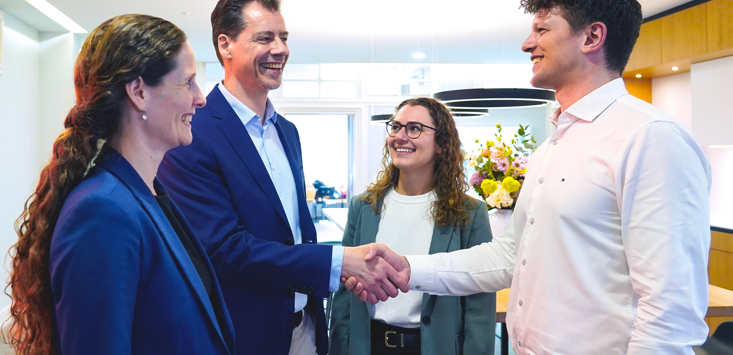 Four professionals in business attire standing in an office. Two women and two men - who are shaking hands smiling.