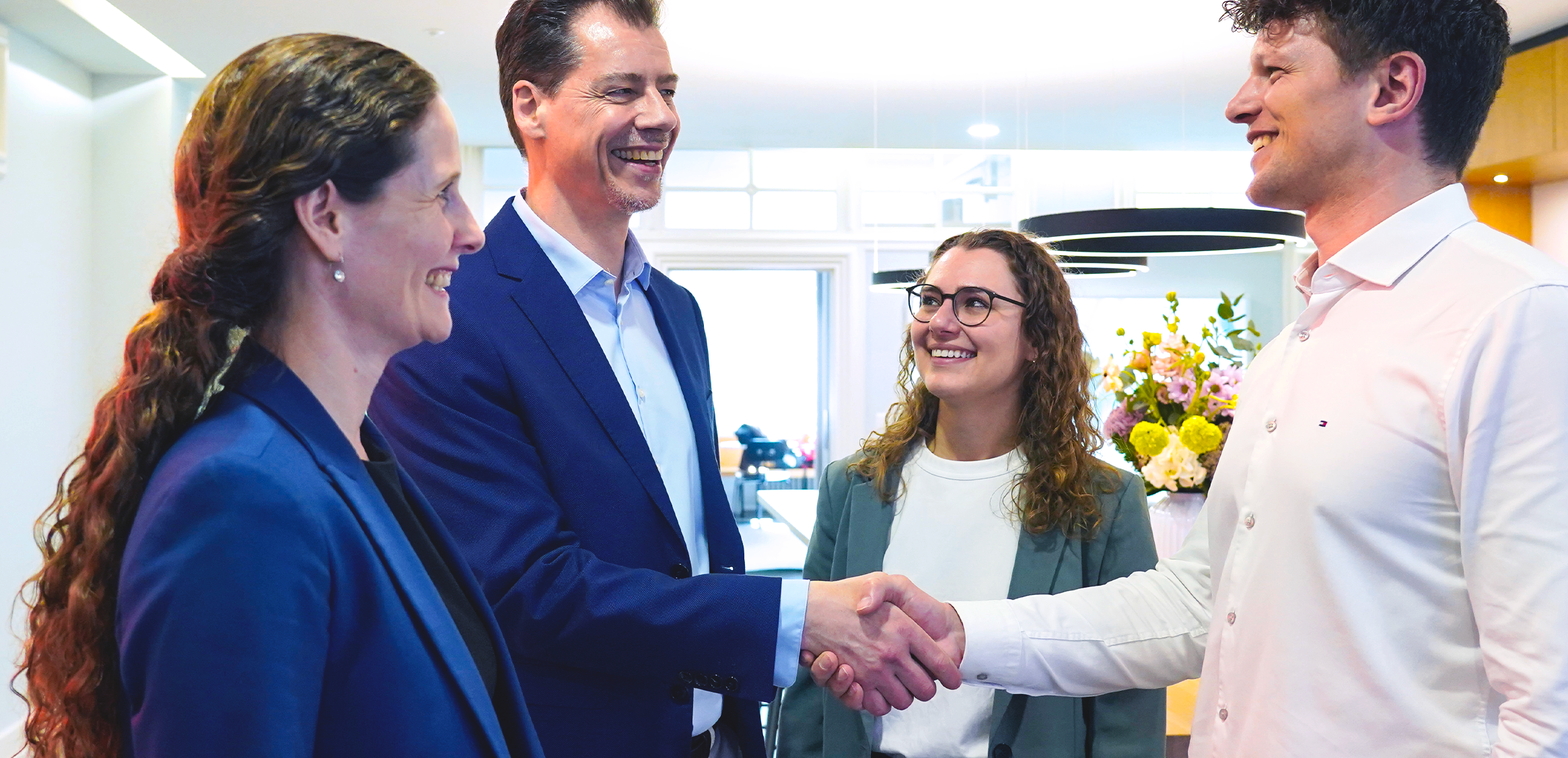 Four professionals in business attire standing in an office. Two women and two men - who are shaking hands smiling.