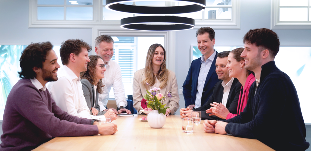 A group of coworkers sit around a conference table in a bright office, smiling and chatting during a meeting. A small vase of flowers sits in the center of the table, with glasses of water in front of several people, while modern circular lights hang overhead.