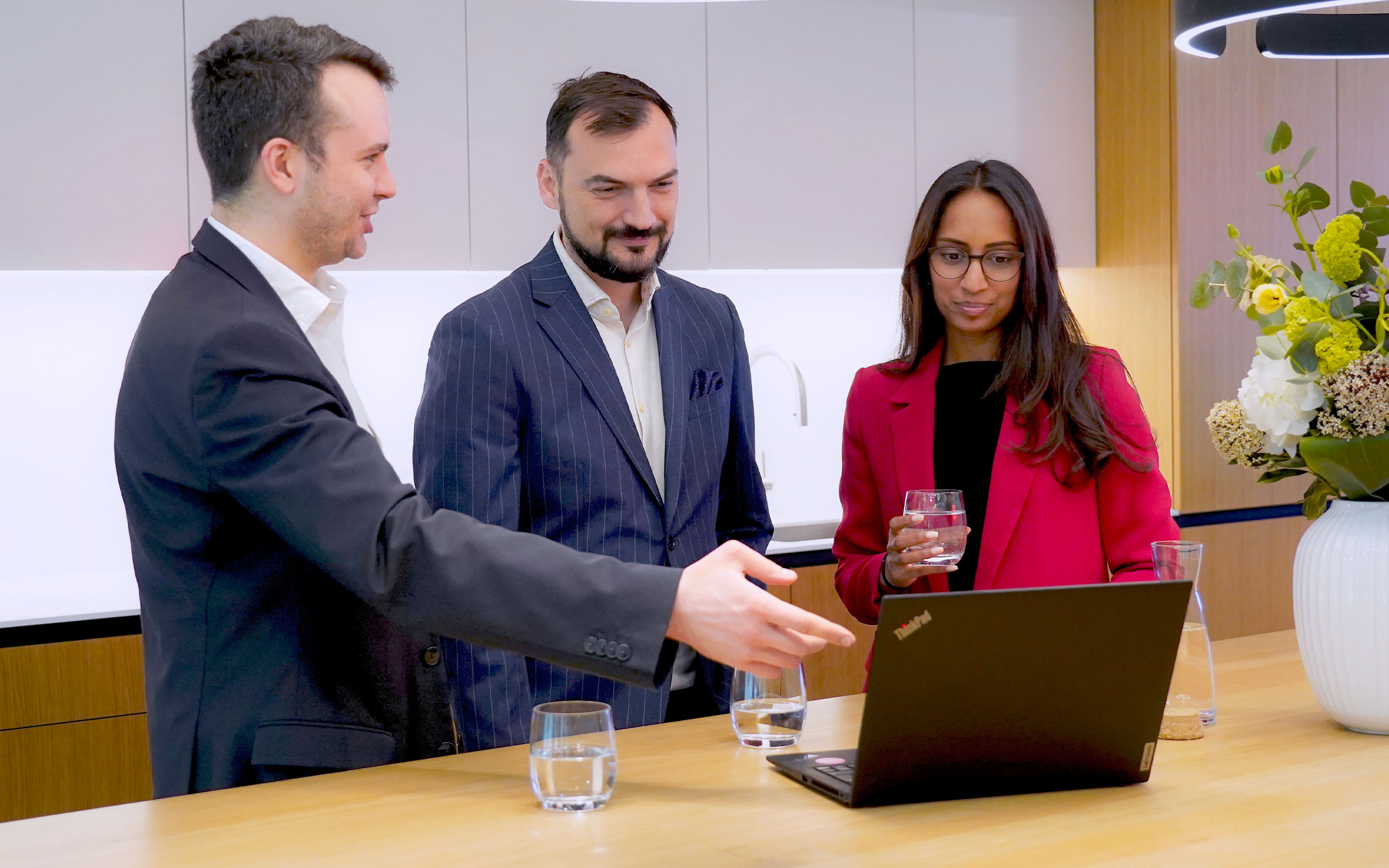 Three professionals in business attire stand around a laptop on a counter in a modern office kitchen. One man gestures toward the screen while speaking, another man looks on with a slight smile, and a woman in a red blazer holds a glass of water and looks at the laptop. Glasses of water and a vase of flowers are visible on the counter.