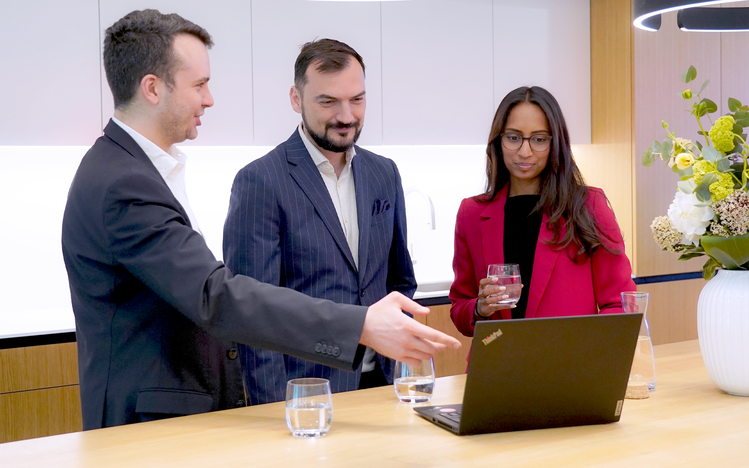 Three professionals in business attire stand around a laptop on a counter in a modern office kitchen. One man gestures toward the screen while speaking, another man looks on with a slight smile, and a woman in a red blazer holds a glass of water and looks at the laptop. Glasses of water and a vase of flowers are visible on the counter.