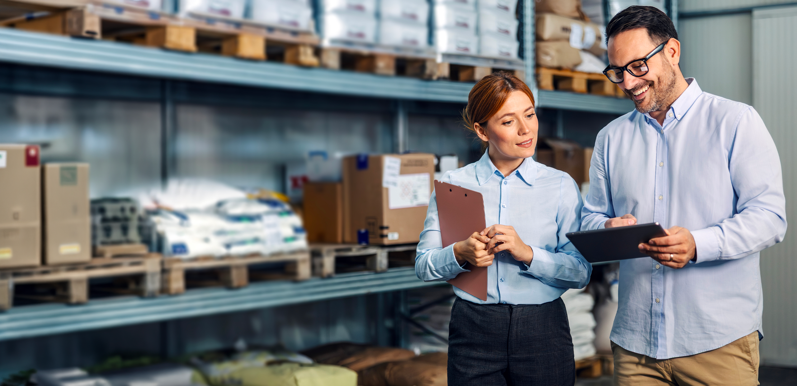 Two retail warehouse employees stand in an aisle of a storage facility, reviewing inventory. One holds a tablet while the other holds a clipboard, and both appear to be discussing stock levels. Shelving units filled with boxes and packaged goods line the background, indicating a logistics or fulfillment environment.