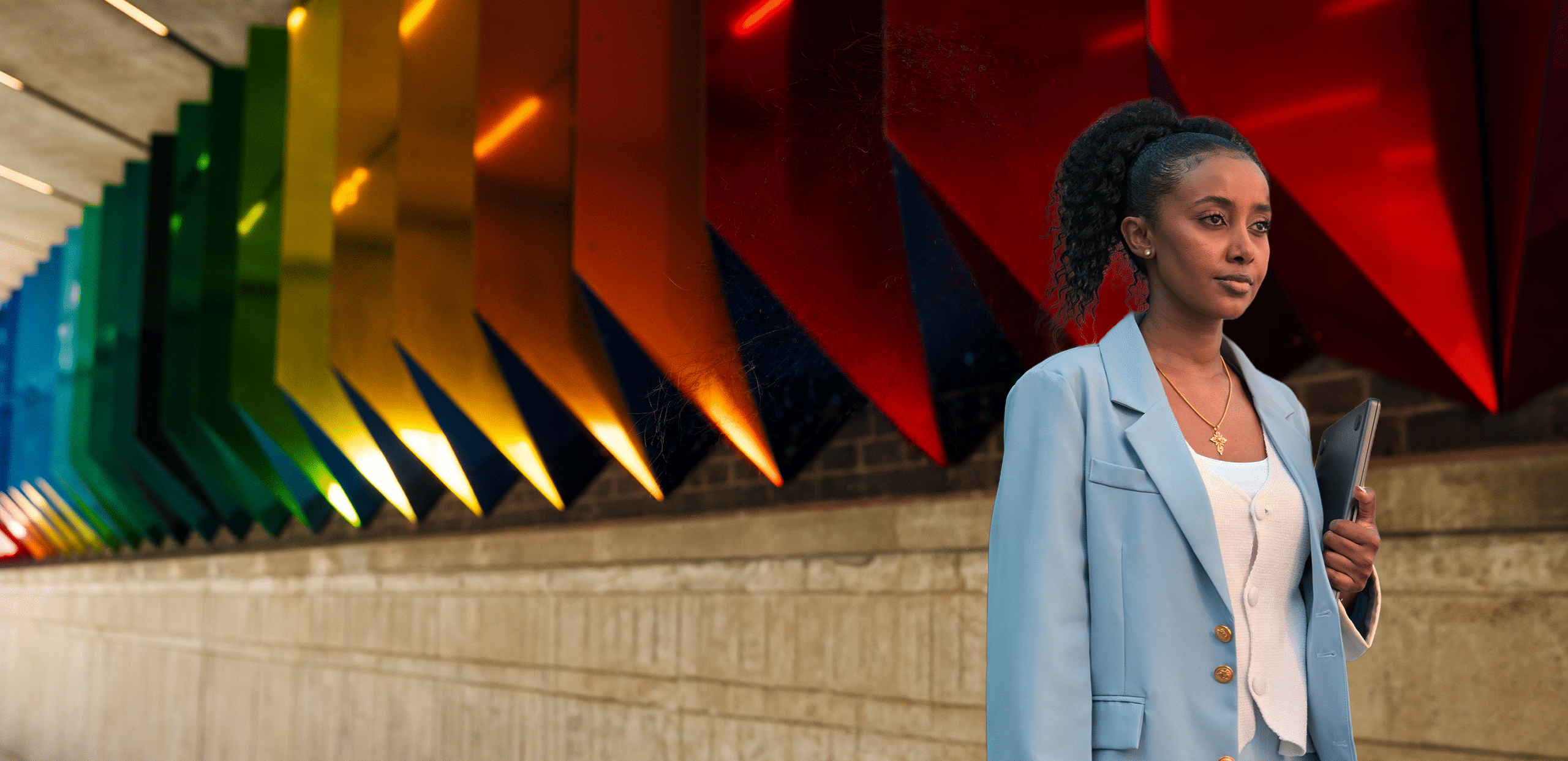 Woman in a light blue blazer walking through a modern architectural corridor with colorful geometric ceiling panels, holding a tablet; a corporate, tech-consulting visual suggesting digital transformation and AI sovereignty.