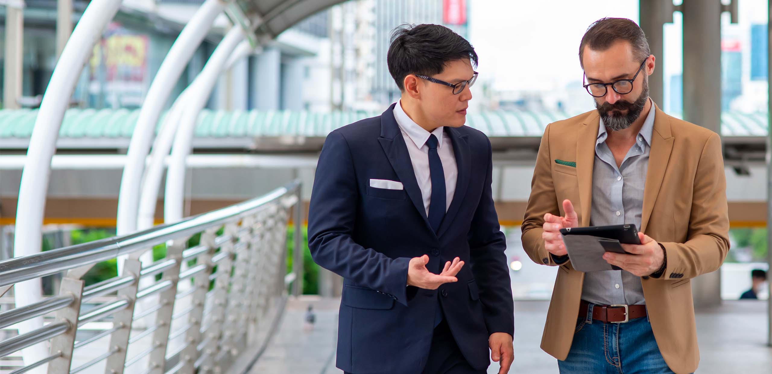 Two businessmen walking together on an urban pedestrian walkway. One wears a navy suit and glasses and gestures as he speaks; the other, bearded, in a tan blazer and jeans, shows him something on a tablet. The scene suggests a telco consultant presenting telecom consolidation approach to a client.