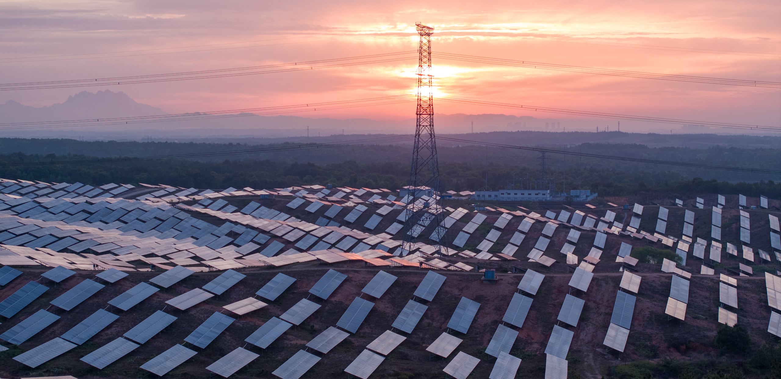 Solar farm on rolling hills at sunset, with a high-voltage power pylon and transmission lines connecting it to the grid. Showcasing connectivity in the energy sector.